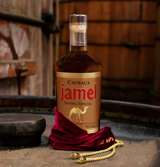 Garrafa elegante de Cachaça Jamel Reserva Especial com rótulo dourado sobre um pano de veludo vermelho e cordão dourado, sugerindo exclusividade e luxo.